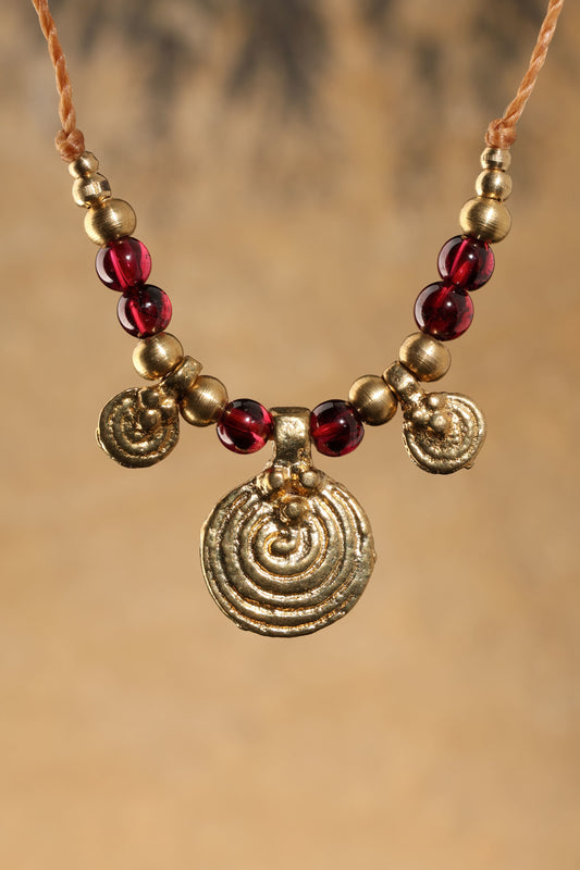 Halskette mit Edelstein- und Messingperlen sowie 3 runden Messinganhängern. Zentraler Messinganhänger mit Spiralsymbol. Kette aus beigem Makrameegarn. Stein und Farbe: Granat –dunkelrot/klar. Für Affirmation: „Ich bin mutig“