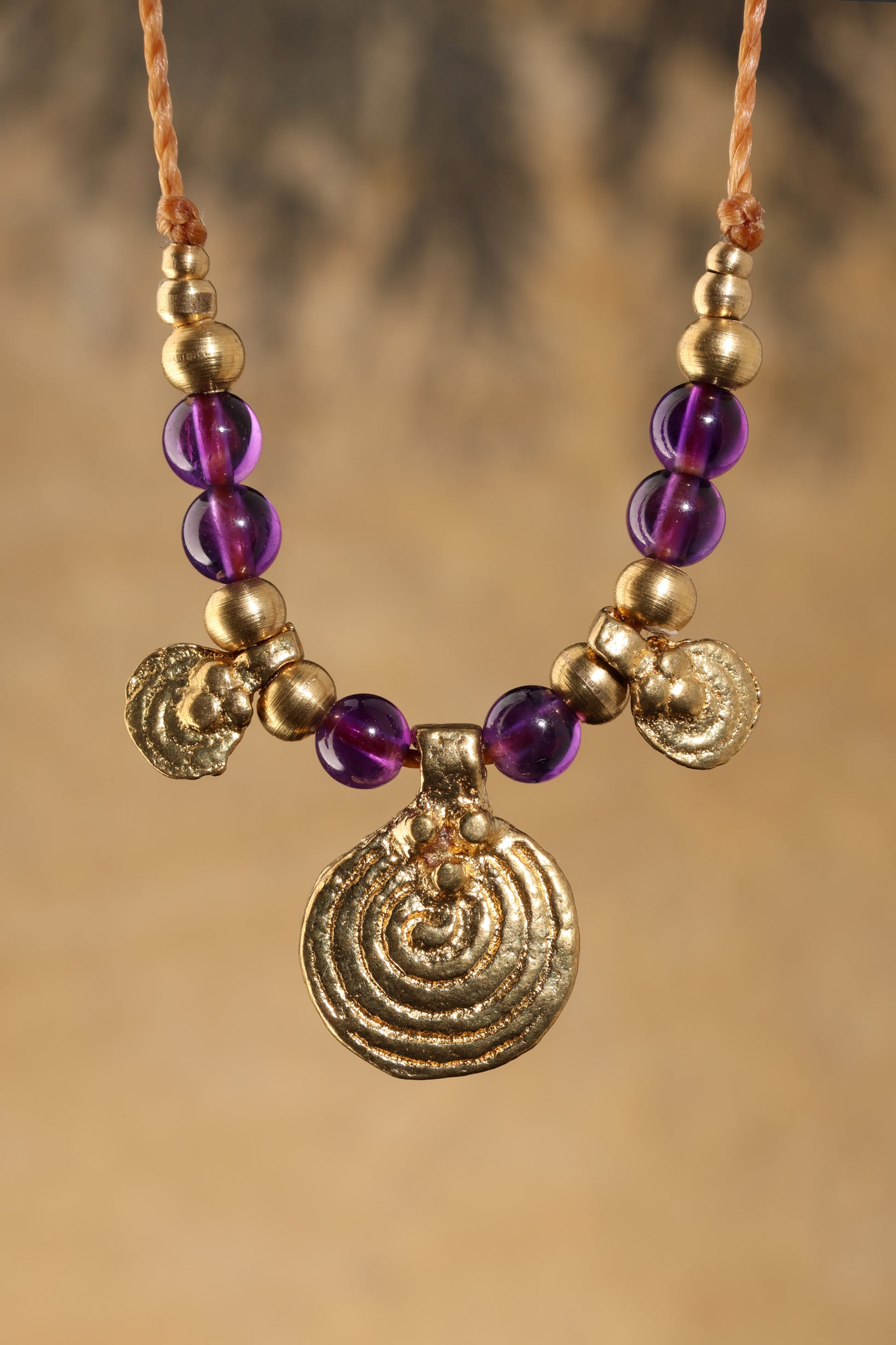 Halskette mit Edelstein- und Messingperlen sowie 3 runden Messinganhängern. Zentraler Messinganhänger mit Spiralsymbol. Kette aus beigem Makrameegarn. Stein und Farbe: Amethyst –lila/klar. Für Affirmation: „Ich bin easy“