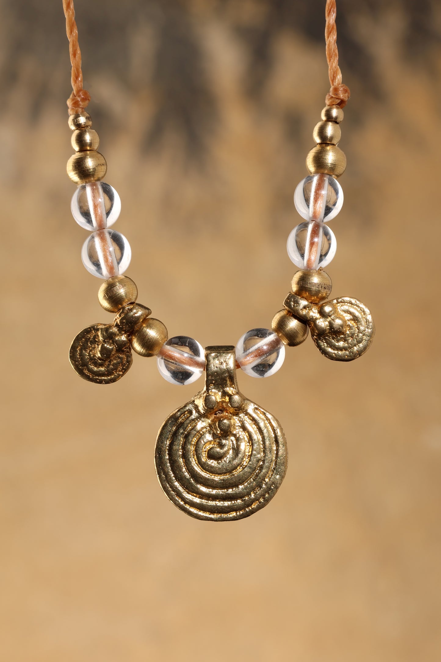 Halskette mit Edelstein- und Messingperlen sowie 3 runden Messinganhängern. Zentraler Messinganhänger mit Spiralsymbol. Kette aus beigem Makrameegarn. Stein und Farbe: Bergkristall –klar. Für Affirmation: „Ich bin im Jetzt“