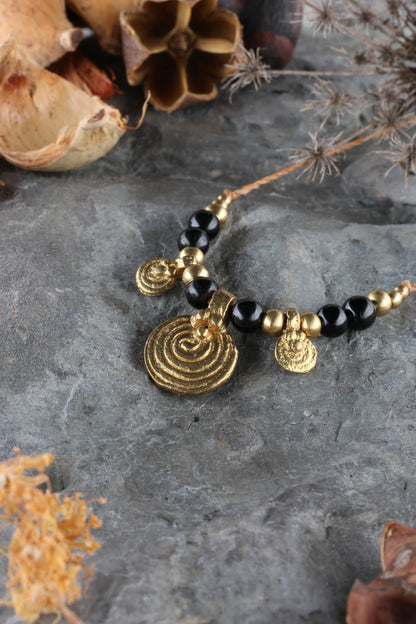 Halskette mit Edelstein- und Messingperlen sowie 3 runden Messinganhängern. Zentraler Messinganhänger mit Spiralsymbol. Kette aus beigem Makrameegarn. Stein und Farbe: Turmalin –schwarz. Für Affirmation: „Ich bin safe“