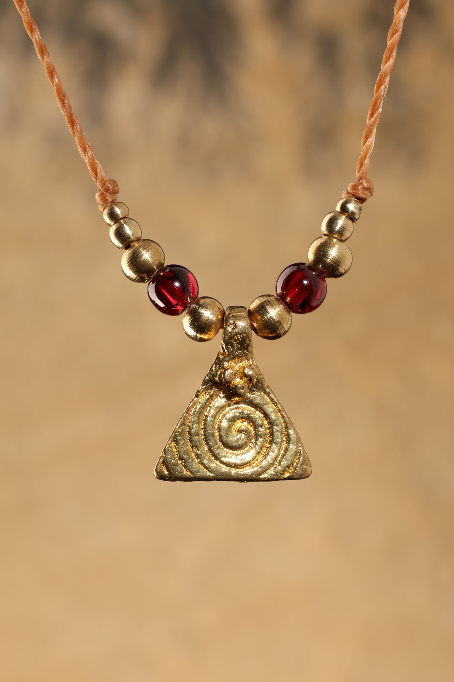 Halskette mit Edelstein- und Messingperlen sowie einem dreieckigen Messinganhänger mit Spiralsymbol. Kette aus beigem Makrameegarn. Stein und Farbe: Granat –dunkelrot/klar. Für Affirmation: „Ich bin mutig“