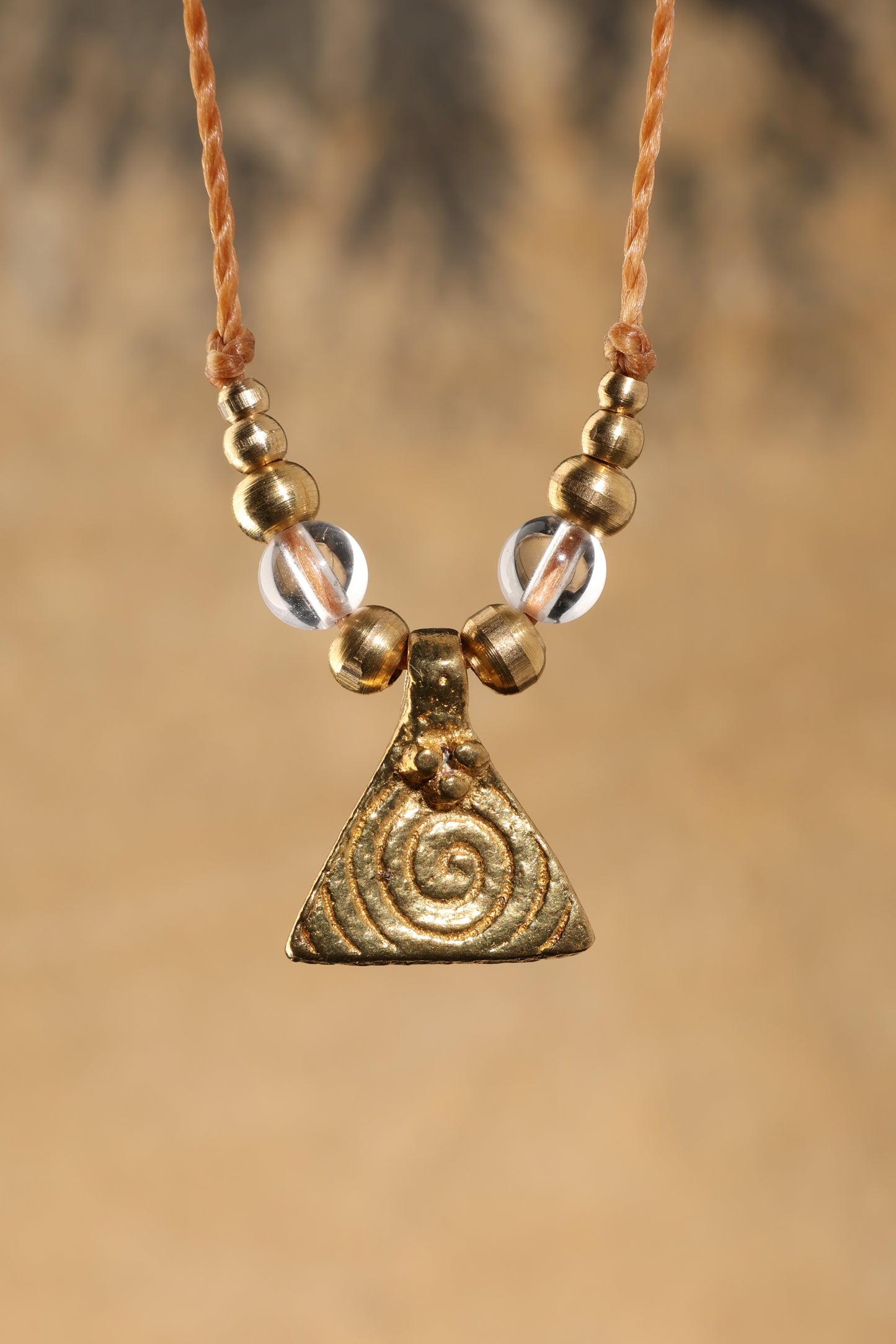 Halskette mit Edelstein- und Messingperlen sowie einem dreieckigen Messinganhänger mit Spiralsymbol. Kette aus beigem Makrameegarn. Stein und Farbe: Bergkristall – klar. Für Affirmation: „Ich bin im Jetzt“