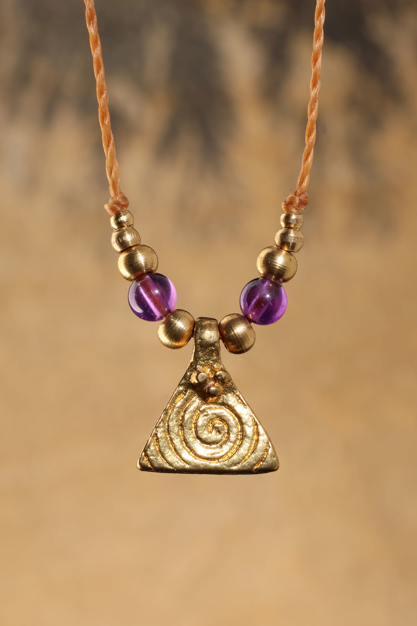 Halskette mit Edelstein- und Messingperlen sowie einem dreieckigen Messinganhänger mit Spiralsymbol. Kette aus beigem Makrameegarn. Stein und Farbe: Amethyst – lila/klar. Für Affirmation: „Ich bin easy“