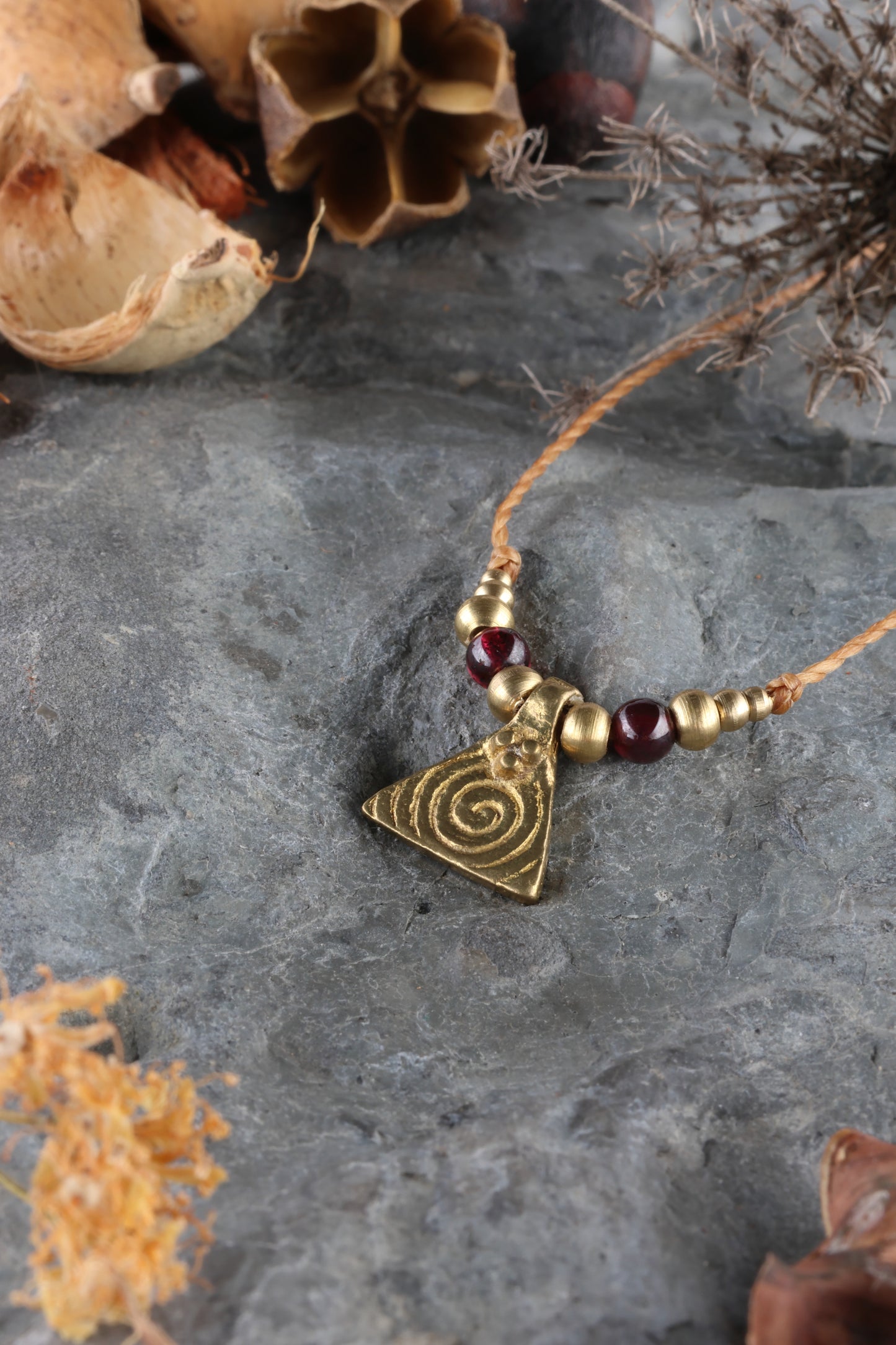 Halskette mit Edelstein- und Messingperlen sowie einem dreieckigen Messinganhänger mit Spiralsymbol. Kette aus beigem Makrameegarn. Stein und Farbe: Granat –dunkelrot/klar. Für Affirmation: „Ich bin mutig“